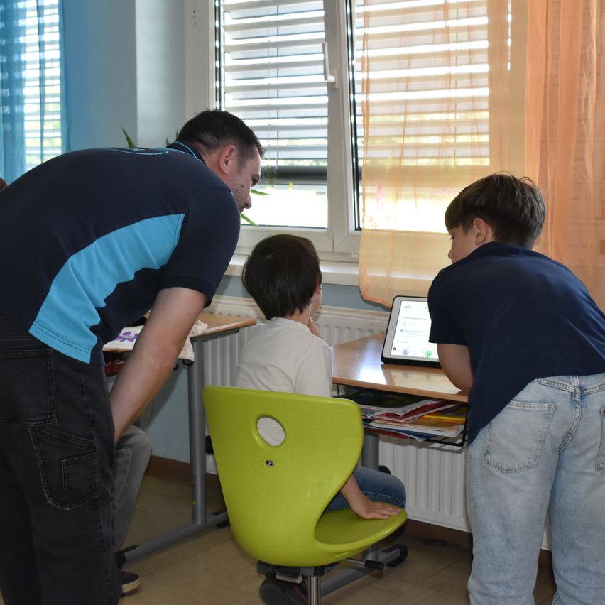 Ein Mann und zwei Jungen schauen sich ein Tablet an. Ein Junge sitzt auf einem grünen Stuhl.