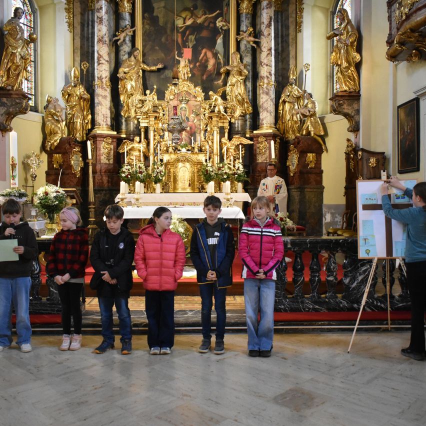 Eine Gruppe von Kindern steht vor einem Kirchenaltar, mit einem Priester im Hintergrund. Der Altar ist mit Blumen und goldenen Statuen geschmückt.