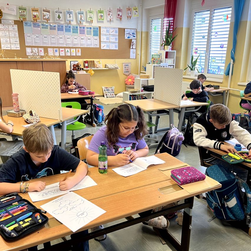 Schüler sitzen in einem Klassenzimmer an Tischen, tragen Masken. Ein Mädchen schreibt, während ein Junge zeichnet. Verschiedene Utensilien liegen auf den Tischen, und Trennwände trennen die Schüler.