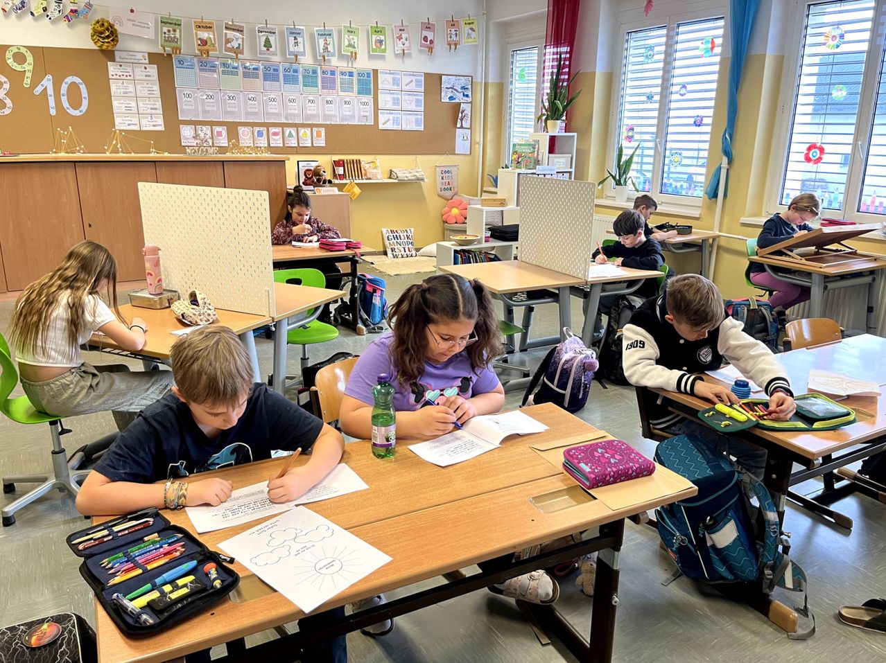 Schüler sitzen in einem Klassenzimmer an Tischen, tragen Masken. Ein Mädchen schreibt, während ein Junge zeichnet. Verschiedene Utensilien liegen auf den Tischen, und Trennwände trennen die Schüler.