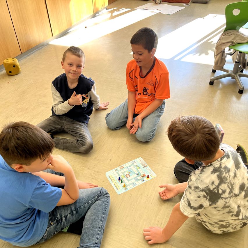 Vier Jungen sitzen auf dem Boden und spielen ein Brettspiel. Einer zeigt auf das Brett, während ein anderer lächelt. Ein gelber Würfel und eine Jacke liegen in der Nähe.
