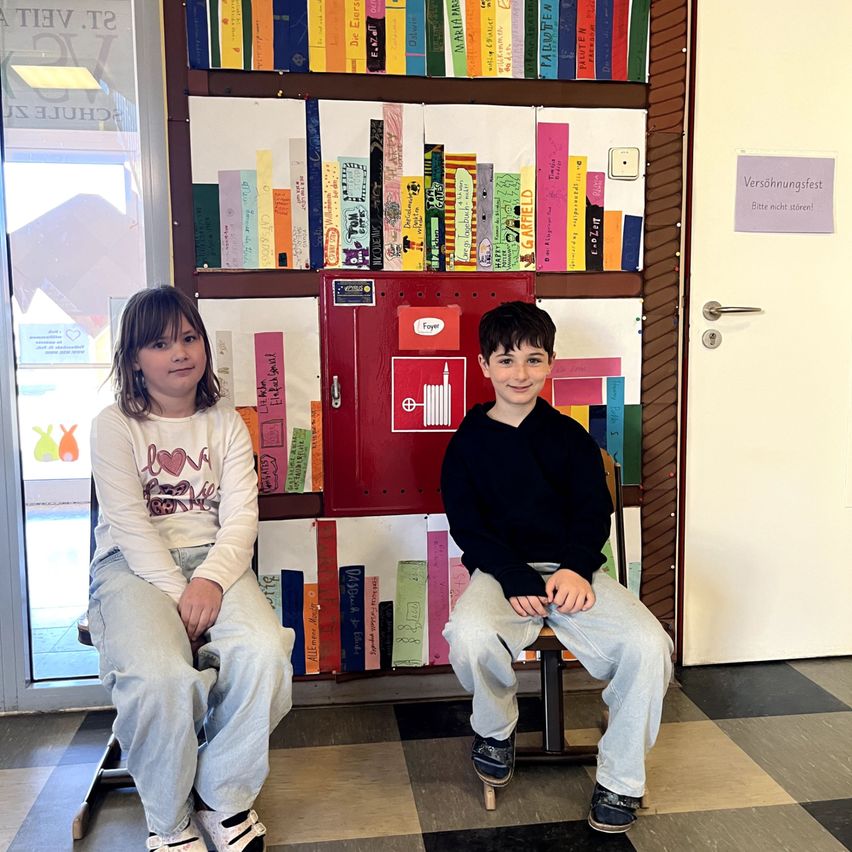 Zwei Kinder sitzen nebeneinander vor einem bunten Regal. Das Regal hat viele Bücher darauf. Der Junge trägt einen schwarzen Pullover.