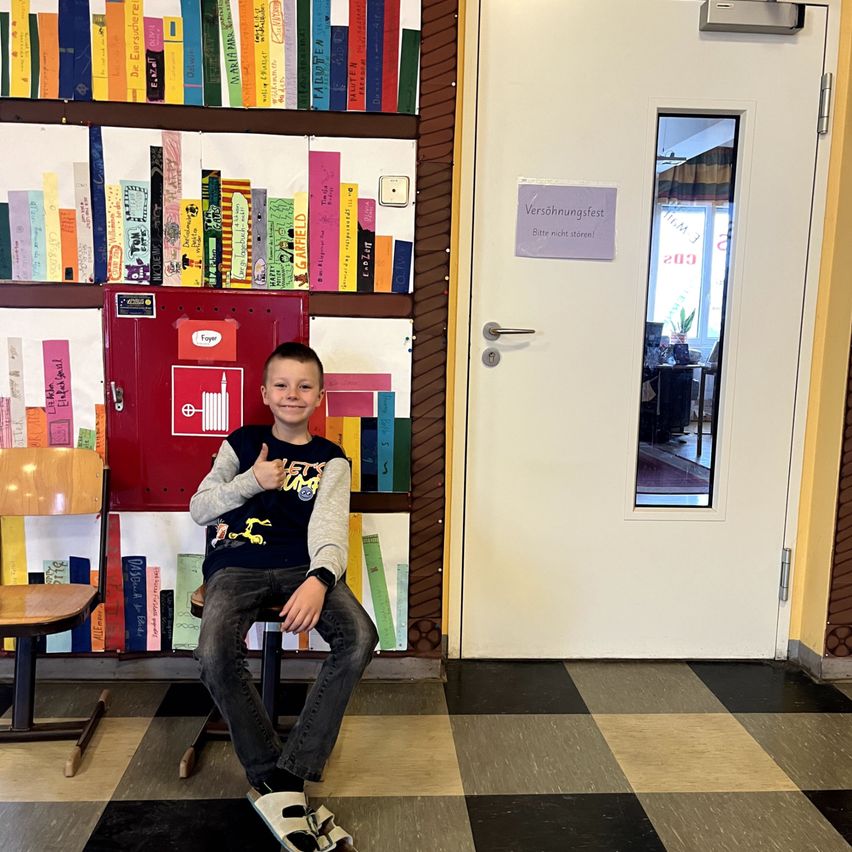 Ein junger Junge sitzt in einem Stuhl in einem Raum mit einem Bücherregal und einer Tür. Ein lila Schild an der Tür steht 'Versöhnungsfest'.