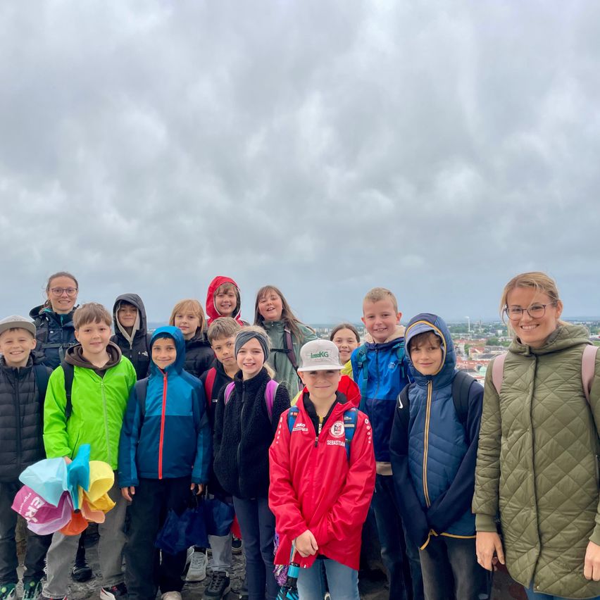 Eine Gruppe von Kindern und Erwachsenen steht auf einem Hügel und trägt verschiedene Jacken und Mützen. Ein Kind hält bunte Hüte, während der Hintergrund einen bewölkten Himmel zeigt.
