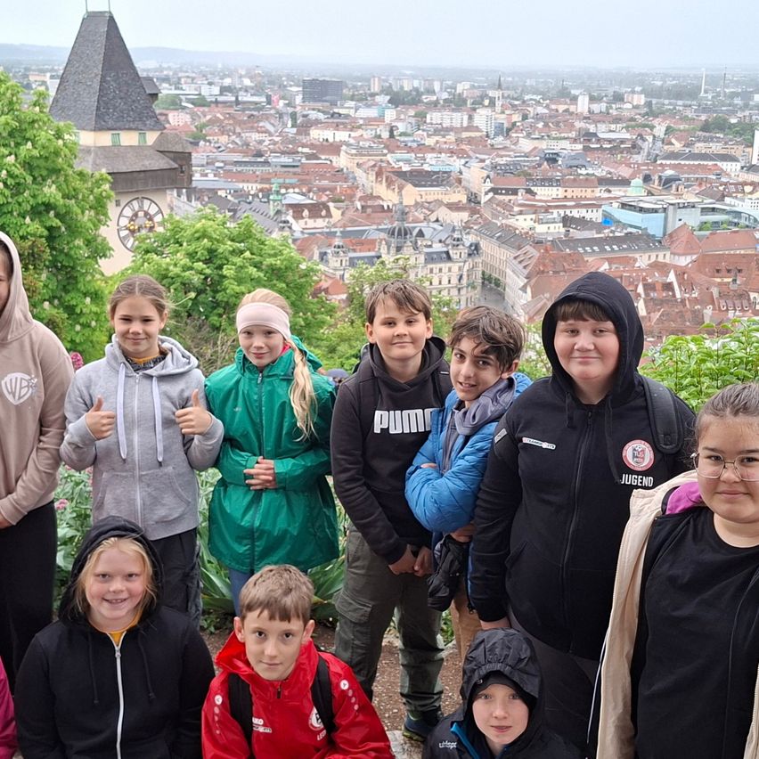 Eine Gruppe von Kindern und Teenagern posiert für ein Foto mit Blick auf die Stadt im Hintergrund. Bäume und ein Uhrturm sind sichtbar.