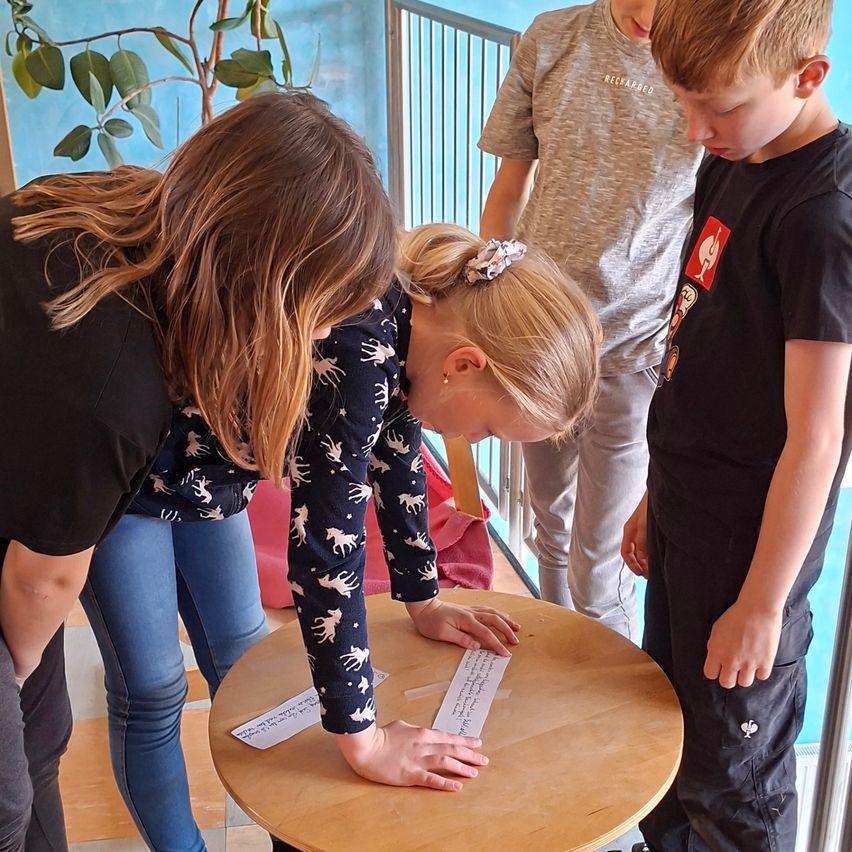 Mehrere Kinder versammeln sich um einen Tisch mit Papieren. Ein Mädchen in Pyjamas mit Hirsch-Aufdruck schaut auf das Papier. Ein Junge in einem schwarzen T-Shirt beobachtet. Ein Erwachsener und ein weiteres Kind stehen hinter ihnen.
