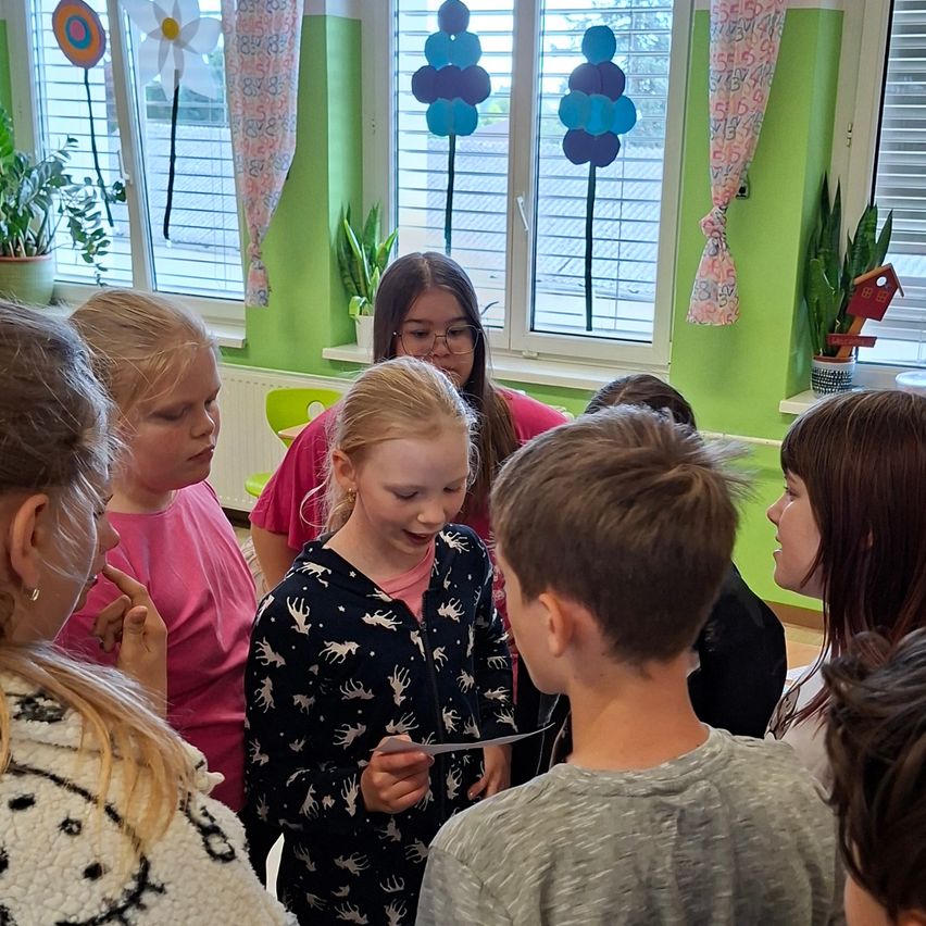 Eine Gruppe von Kindern ist drinnen versammelt, einige unterhalten sich. Ein Mädchen mit Brille hält ein Stück Papier in der Hand. Der Raum hat grüne Wände und weiße Vorhänge.