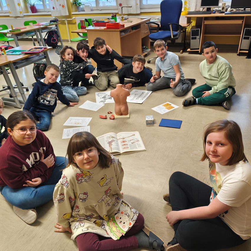 Eine Gruppe von Schülern sitzt in einem Klassenzimmer in einem Kreis um eine Puppe. Papiere, Bücher und andere Gegenstände sind auf dem Boden verstreut.