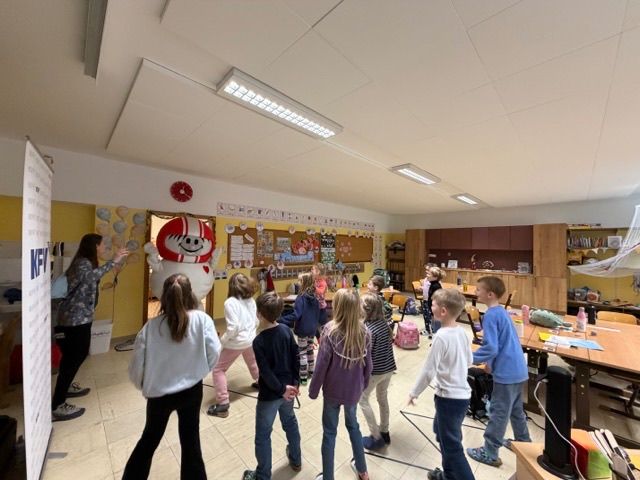 Ein Klassenzimmer mit Kindern und einer Maskottchen; eine Frau steht neben einer Tür mit einem Maskottchen neben ihr.