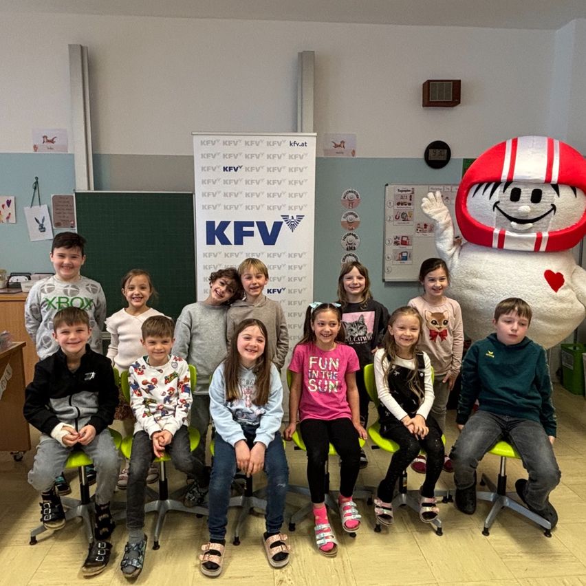 Eine Gruppe von Kindern sitzt in einem Klassenzimmer, lächelt und posiert für ein Foto. Sie tragen Winterkleidung und Turnschuhe. Ein Maskottchen mit rotem Helm und Herz steht hinter ihnen. Eine Tafel mit Text ist an der Wand angebracht.