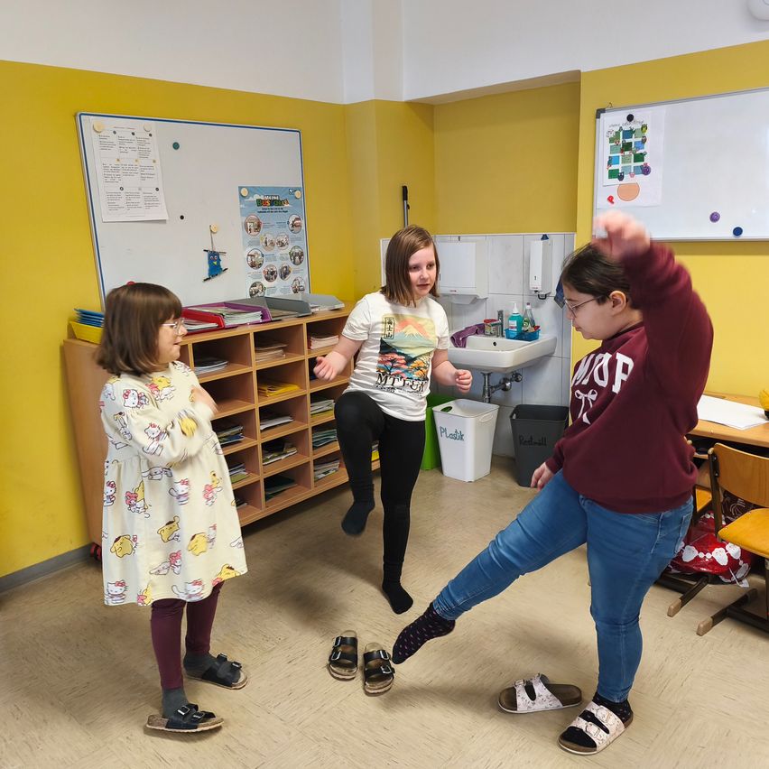 Drei Mädchen in einem Klassenzimmer spielen ein Spiel, ein Mädchen springt, ein anderes schaut zu und das dritte steht in der Nähe des Waschbeckens.