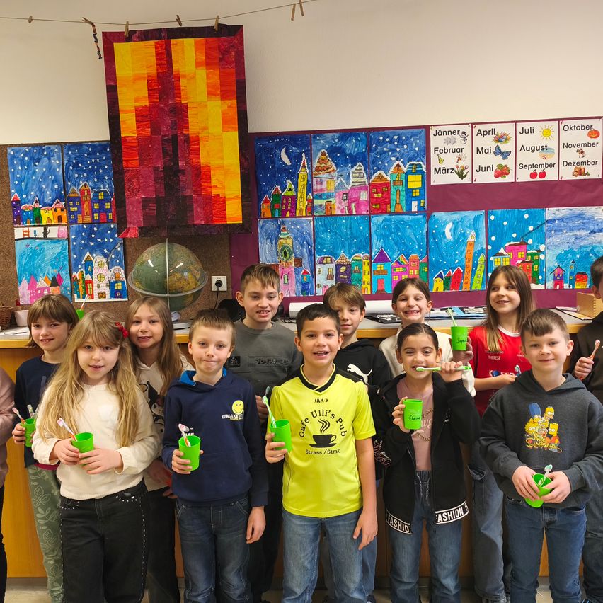 Eine Gruppe von Kindern in einem Klassenzimmer mit einem Globus und Gemälden im Hintergrund, die grüne Becher und Zahnbürsten halten.