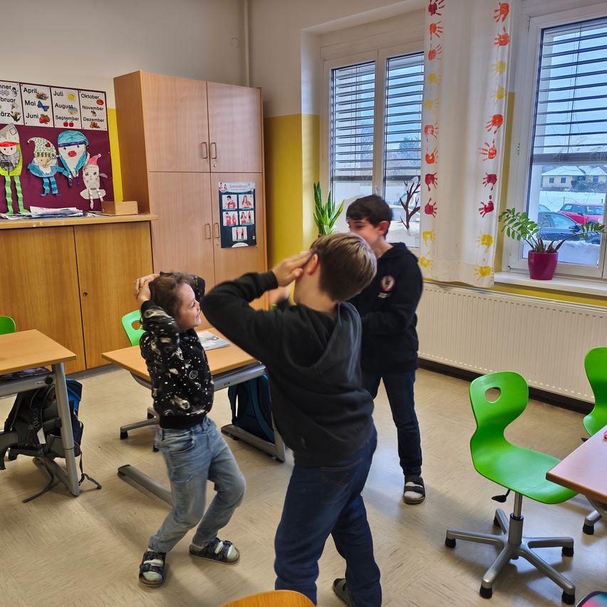 Drei Jungen in einem Klassenzimmer beschäftigen sich mit einer verspielten Aktivität. Ein Junge scheint einen anderen spielerisch auf den Kopf zu schlagen.