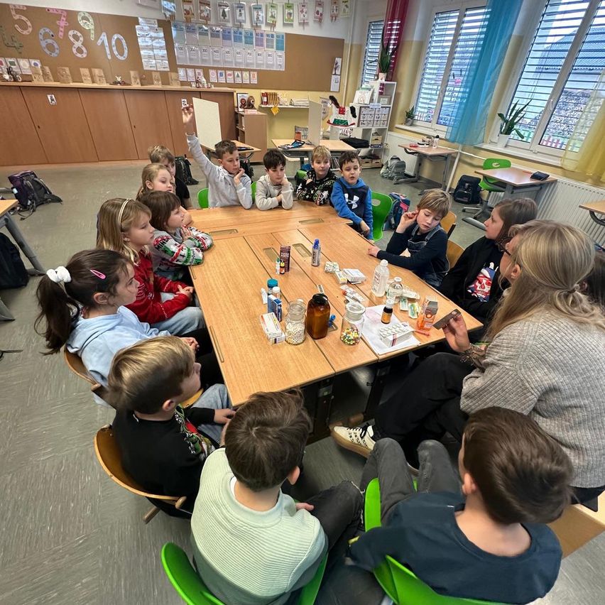 Ein Klassenzimmer mit Kindern und einer Lehrerin, die um einen Tisch herum sitzen, auf dem verschiedene Gegenstände stehen. Die Kinder sind aufmerksam, und die Lehrerin erklärt etwas.