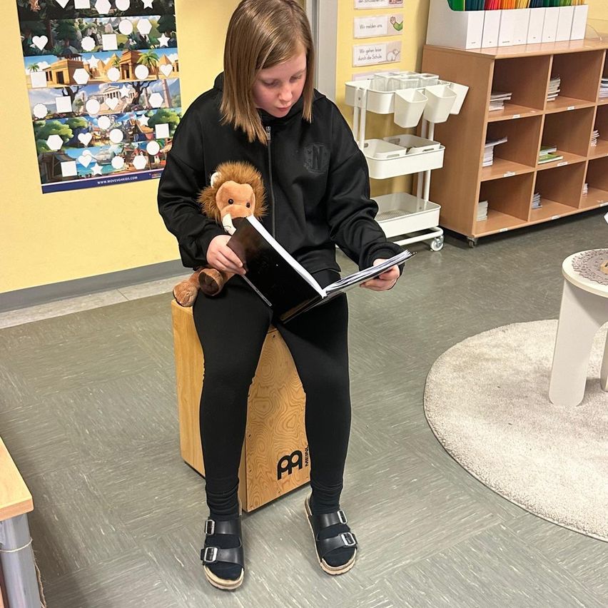 Ein junges Mädchen sitzt in einem Klassenzimmer auf einem Holzhocker und liest ein Buch, während es einen weichen Löwen hält.