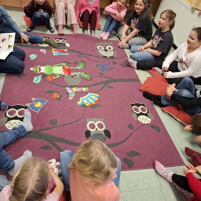 Eine Gruppe von Kindern sitzt im Kreis auf einem violetten Teppich mit Eulen, liest ein Buch und singt. Sie tragen verschiedene Kleidungsstücke und Schuhe.