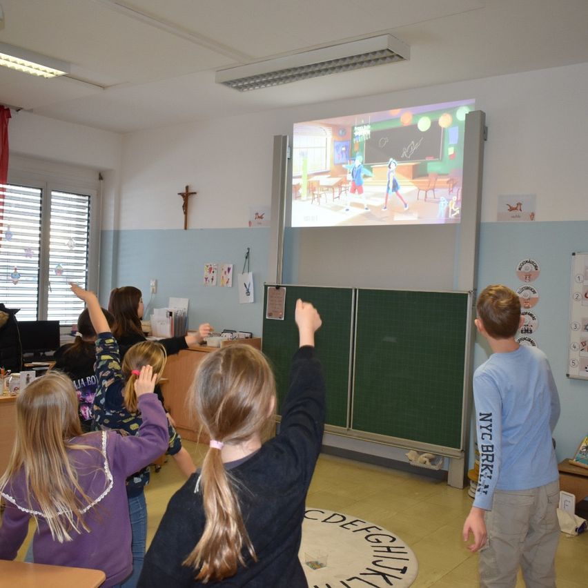 Ein Klassenzimmer mit Schülern, die einen animierten Film auf einer Projektionsleinwand ansehen, eine Mädchen erhebt die Hand und ein Junge vor einer Tafel.