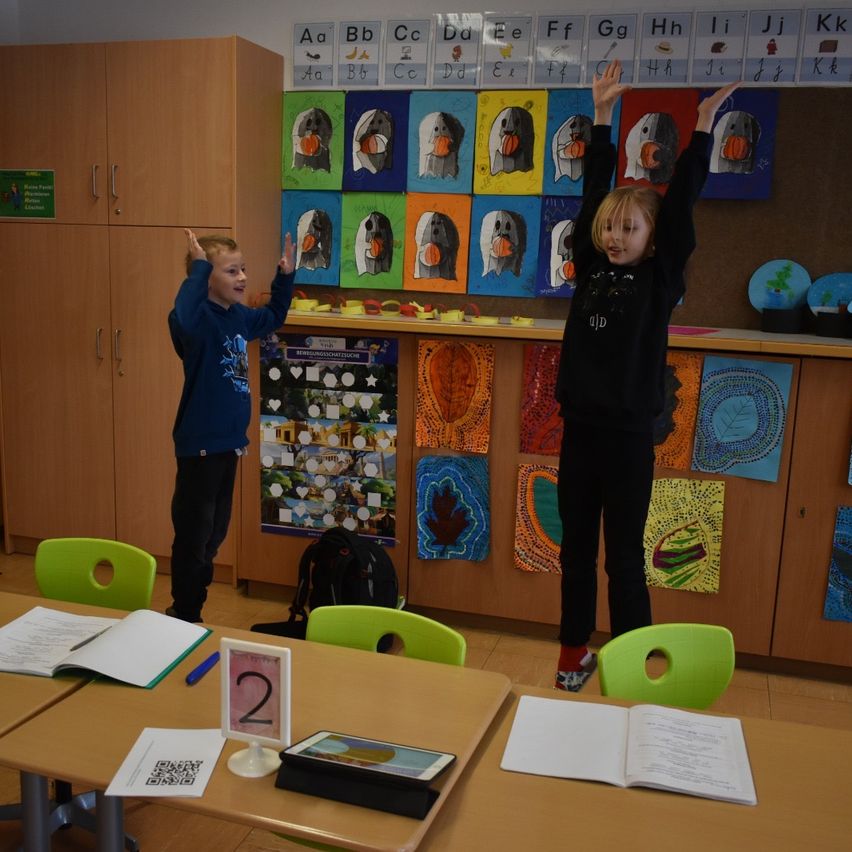 Zwei Kinder stehen in einem Klassenzimmer mit erhobenen Händen. Hinter ihnen ist eine Wand mit Alphabet-Postern und Kunst. Ein Tisch davor hat Bücher, ein Tablet und ein Nummernschild.