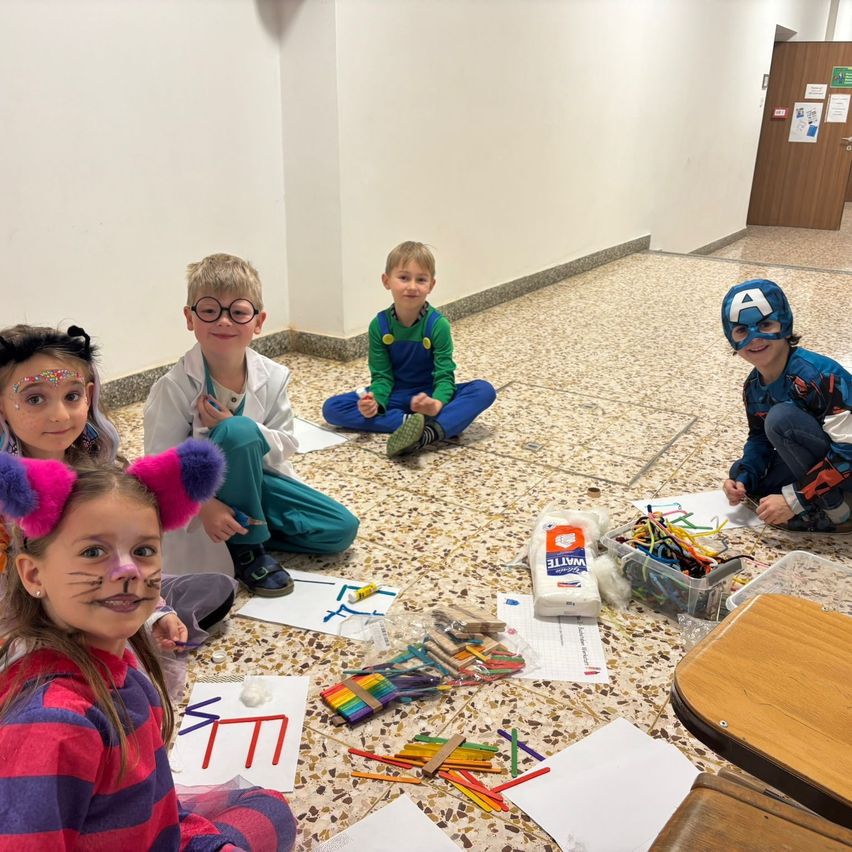 Fünf Kinder sitzen auf dem Boden und sind in einer Bastelaktivität beschäftigt. Sie haben Gesichtsbemalung und tragen Kostüme. Ein Kind ist als Wissenschaftler verkleidet.