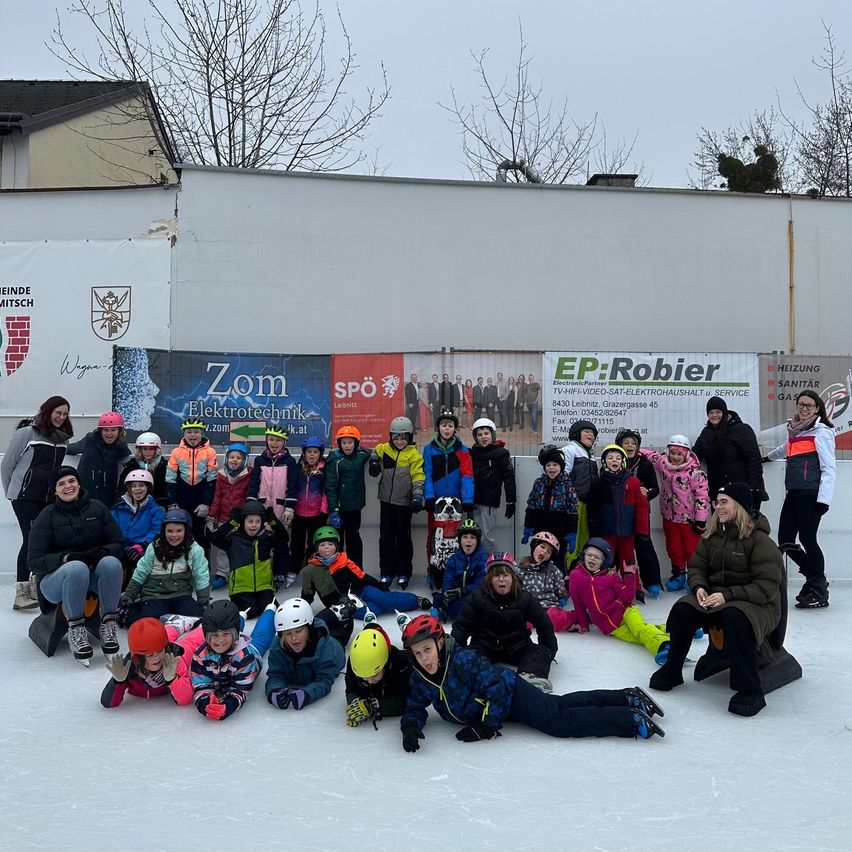 Eine Gruppe von Kindern und Erwachsenen posiert für ein Foto auf einer Eisbahn, trägt Winterkleidung und Helme. Sie stehen vor einer Wand mit verschiedenen Bannern, darunter eines für Elektrotechnik.