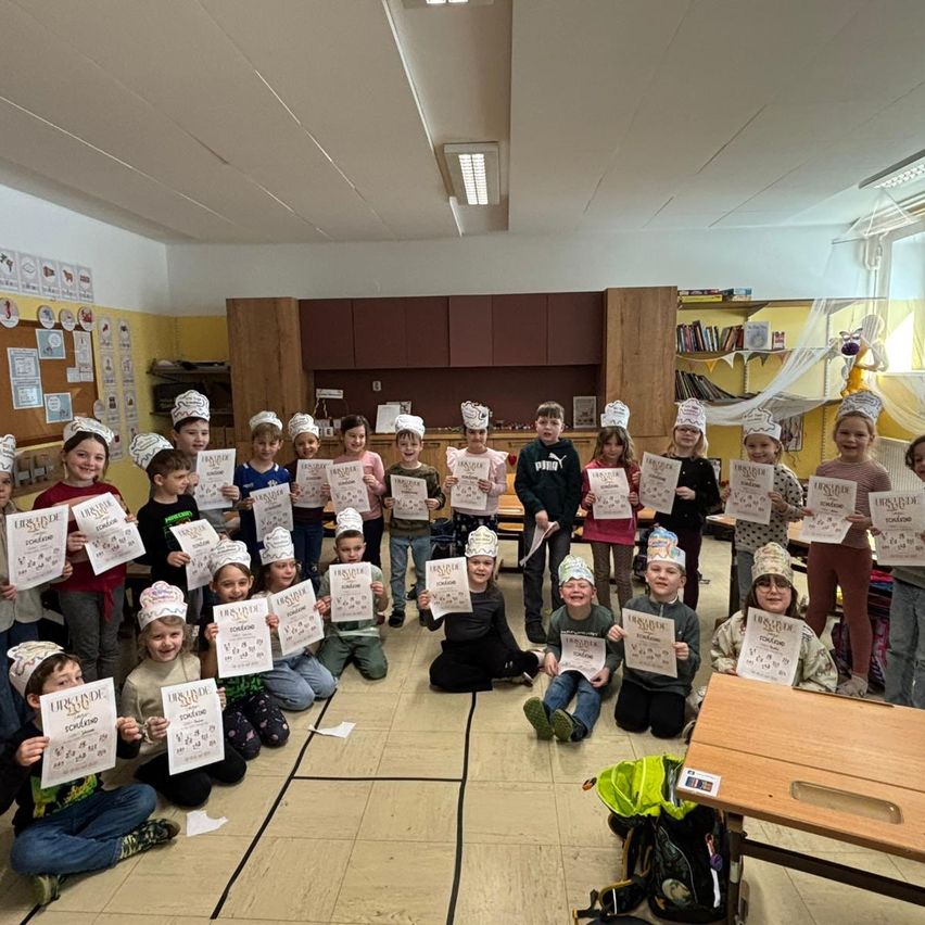 Ein Klassenzimmer voller Kinder mit Kochmützen und Urkunden, einige sitzend, andere stehend, in einem Raum mit Schränken und Regalen.