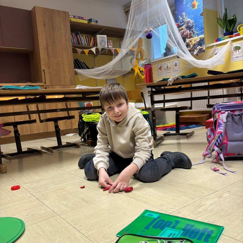 Ein junger Junge sitzt auf dem Boden in einem Klassenzimmer, lächelt und spielt mit roten Objekten auf einem grünen Teppich. Hinter ihm sind Tische, Stühle, ein Rucksack und ein Netz. Der Raum hat eine gelbe Wand mit einem Korallenriff-Bild und Büchern auf einem Regal.
