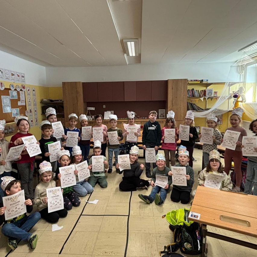 Eine Gruppe von Kindern in einem Klassenzimmer posiert für ein Foto, während sie Kochmützen tragen und Urkunden halten. Sie sitzen und knien auf dem Boden. Einige stehen hinter einem Tisch. Der Raum hat einen Holzschreibtisch, Regale mit Büchern und eine Anschlagtafel.