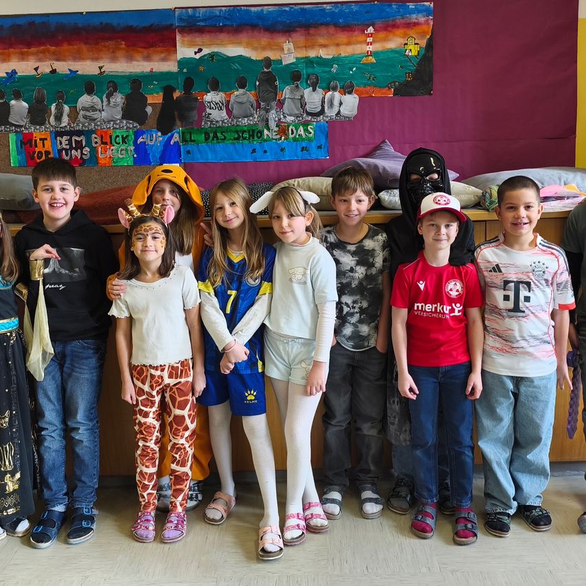 Eine Gruppe von Kindern in Kostümen posiert für ein Foto in einem Klassenzimmer mit einem Wandbild im Hintergrund.
