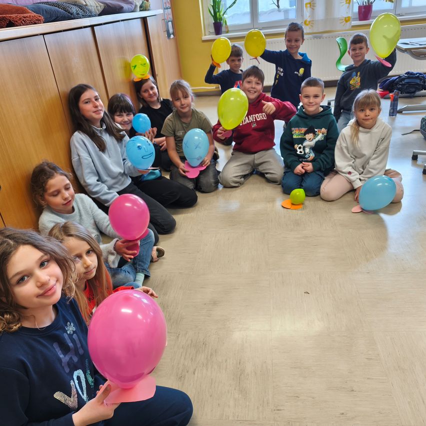 Eine Gruppe von Kindern sitzt im Kreis und hält Luftballons, einige haben Gesichter darauf gezeichnet. Sie lächeln und scheinen Spaß zu haben.