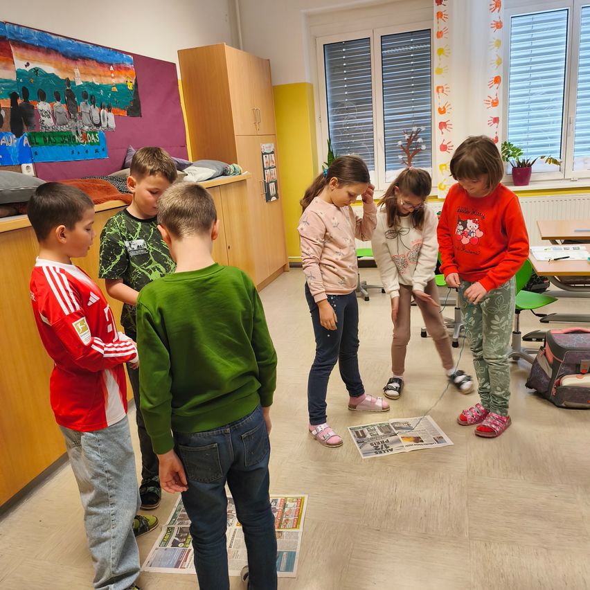 Mehrere Kinder stehen in einem Klassenzimmer mit Zeitungen auf dem Boden, eines hält eine Schnur. Ein Mädchen in Rosa schaut auf das Papier, während ein anderes Mädchen in Grün beobachtet.