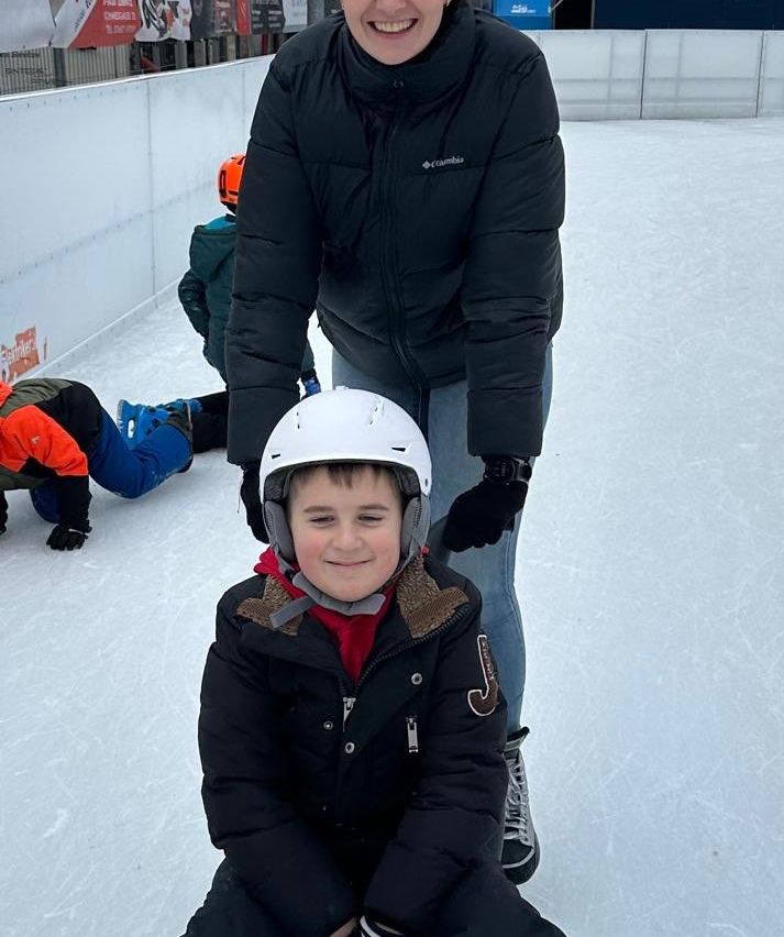 Ein junger Junge mit Helm sitzt lächelnd auf der Eisbahn, mit einer Person hinter ihm. Ein anderes Kind liegt auf dem Eis.