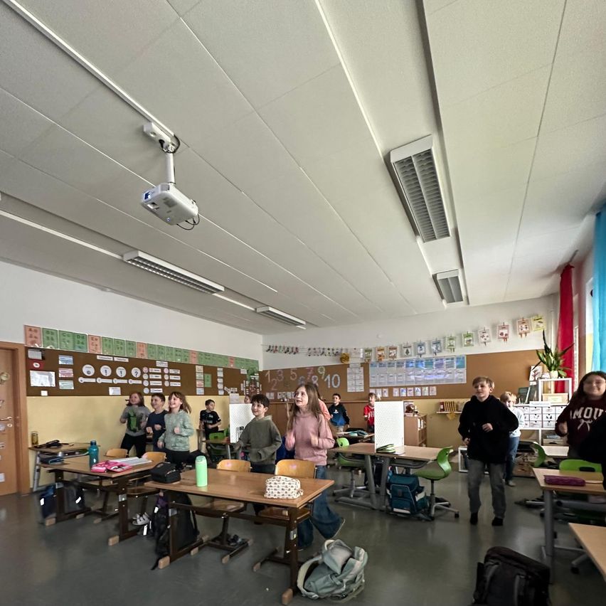 In einem Klassenzimmer sind Schüler versammelt. Einige gehen, andere stehen in der Nähe von Tischen. An der Decke ist ein Beamer montiert, und die Wände sind mit verschiedenen Plakaten und Dekorationen geschmückt.