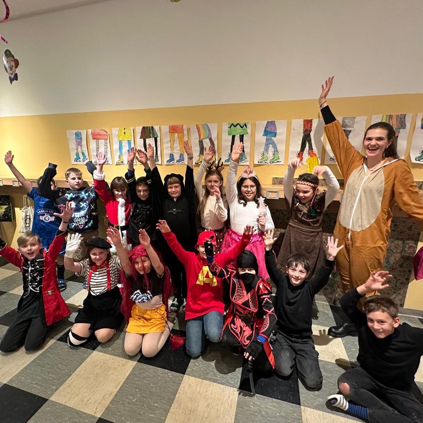 Eine Gruppe von Kindern in verschiedenen Kostümen posiert für ein Foto in einem Klassenzimmer mit einem karierten Boden und gelben Wänden, die mit Zeichnungen verziert sind.