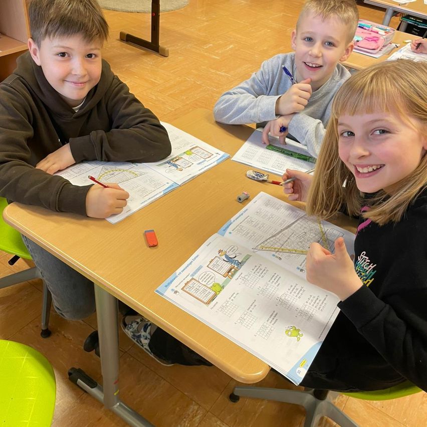 Drei Kinder sitzen an einem Tisch in einem Klassenzimmer. Sie lächeln und arbeiten an ihren Schulaufgaben. Sie halten Stifte in der Hand und haben Papiere vor sich.