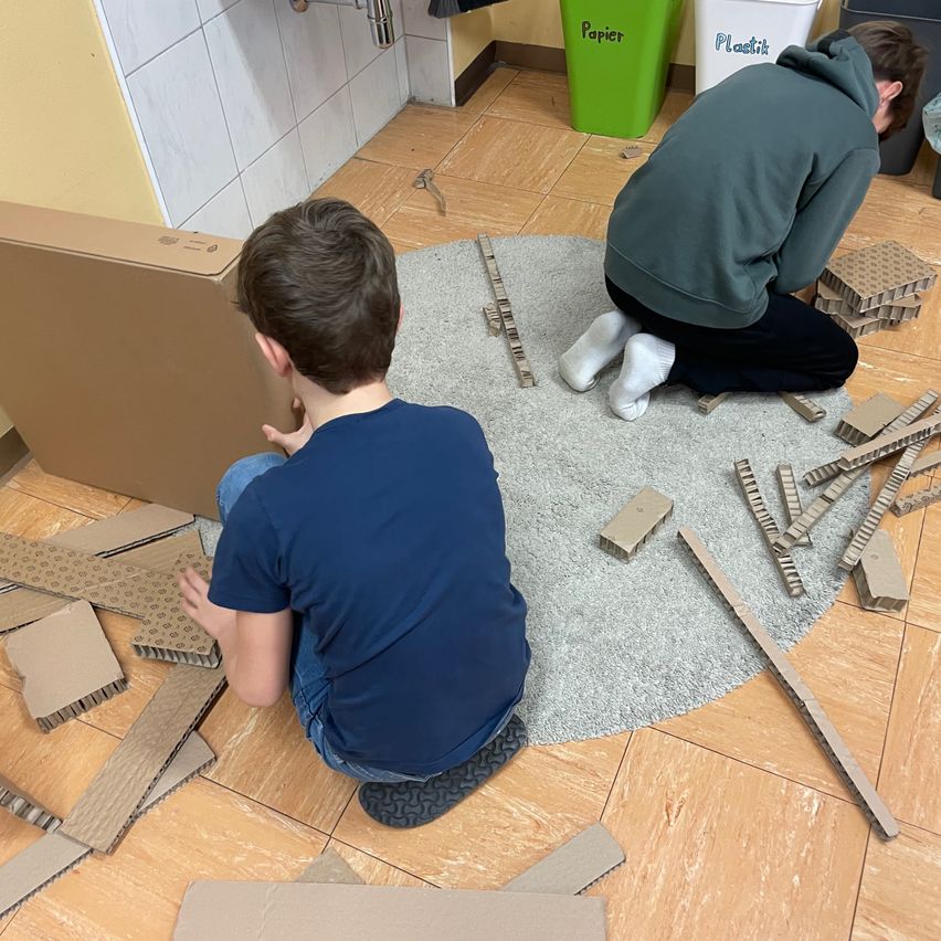 Zwei Jungen sitzen auf dem Boden in einem Raum, einer trägt ein blaues Shirt und der andere eine grüne Kapuzenjacke. Sie sind von verstreuten Pappstücken umgeben. Der Boden ist gefliest, und ein Karton liegt in der Nähe. Ein Mülleimer mit dem Wort 'Papier' ist sichtbar.