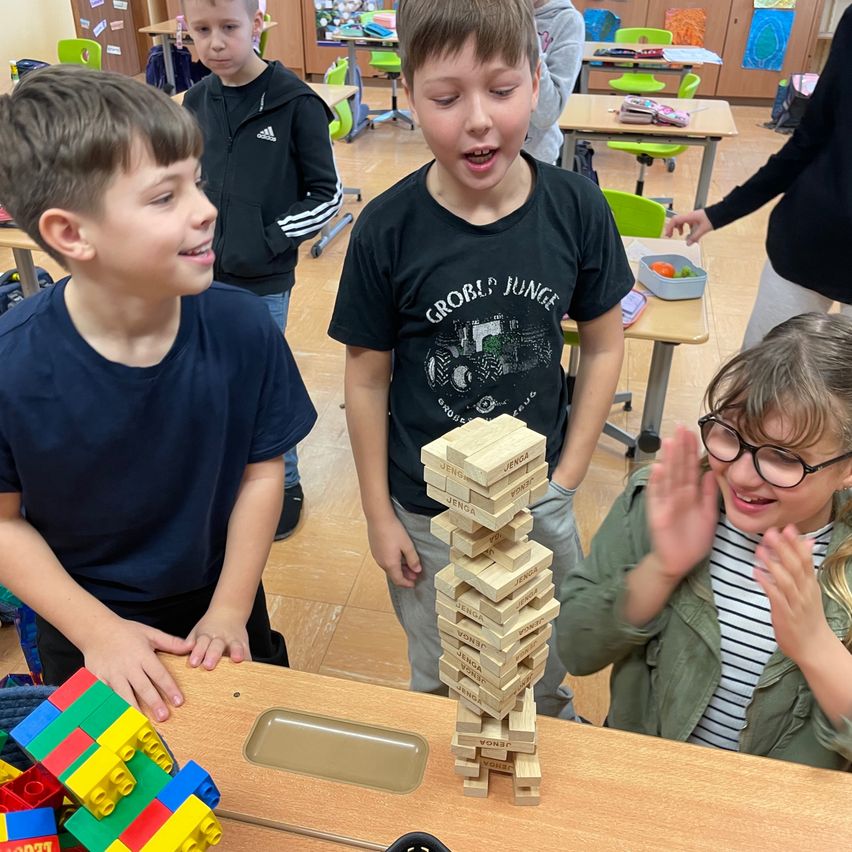 Drei Jungen spielen mit einem Jenga-Turm im Klassenzimmer. Ein Junge schaut zu, ein anderer sieht ihn an und ein Mädchen klatscht.