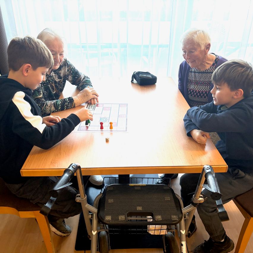 Zwei ältere Frauen und zwei Jungen sitzen um einen Tisch herum und spielen ein Brettspiel mit Würfeln. Ein Junge schaut die Frau an, während der andere spielt.