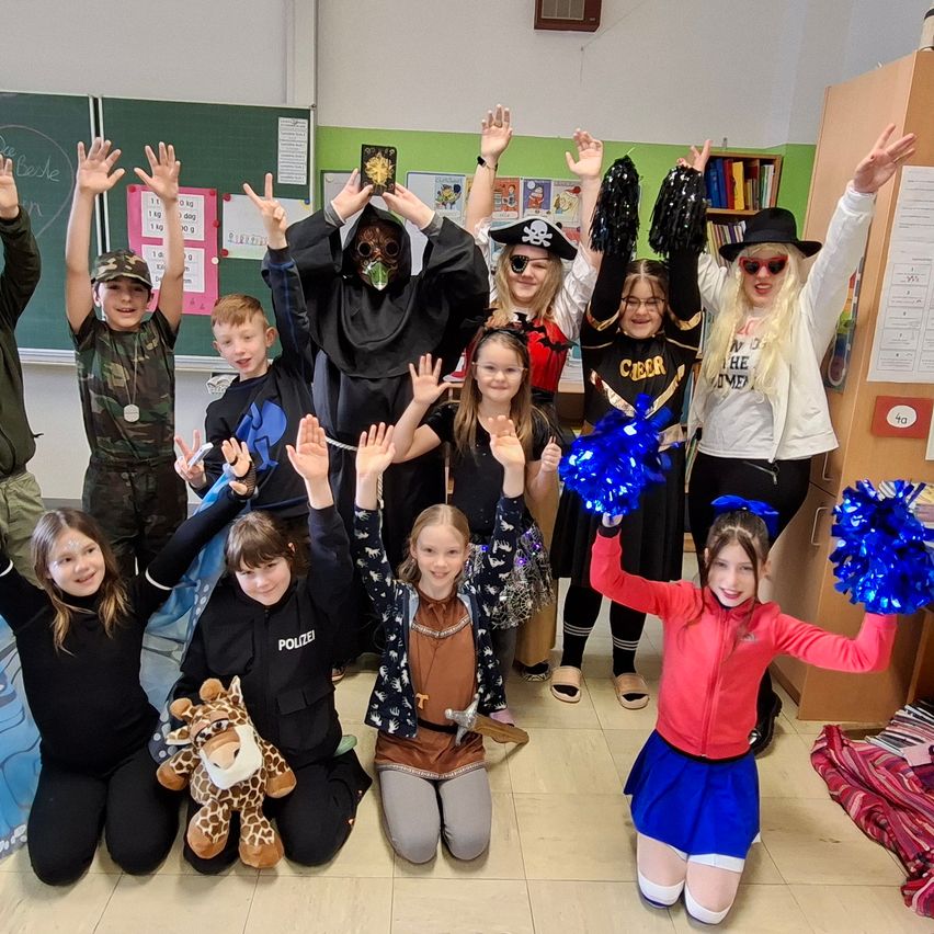 Eine Gruppe von Kindern in verschiedenen Kostümen, darunter Pirat, Hexe und Polizist, posiert für ein Foto im Klassenzimmer. Einige halten blaue Federn.