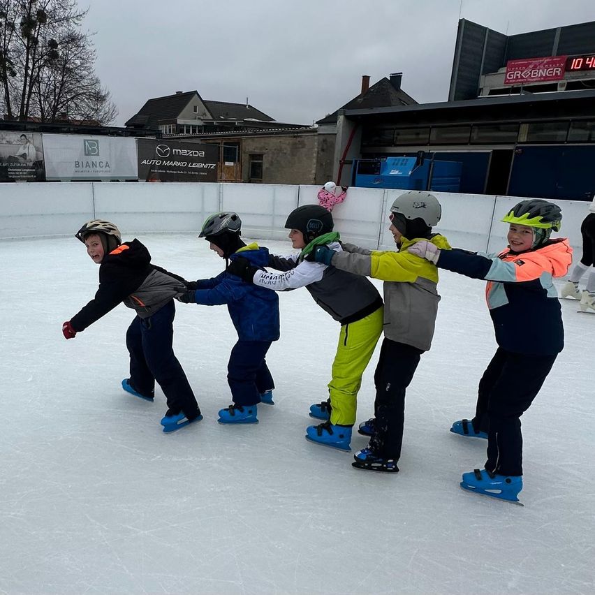 Eine Gruppe junger Leute, die zusammen auf einer Freiluft-Eisbahn Schlittschuh laufen, alle mit Helmen und bunten Jacken. Hinter ihnen steht ein Gebäude mit Schildern und Bannern und eine verschneite Landschaft im Hintergrund.