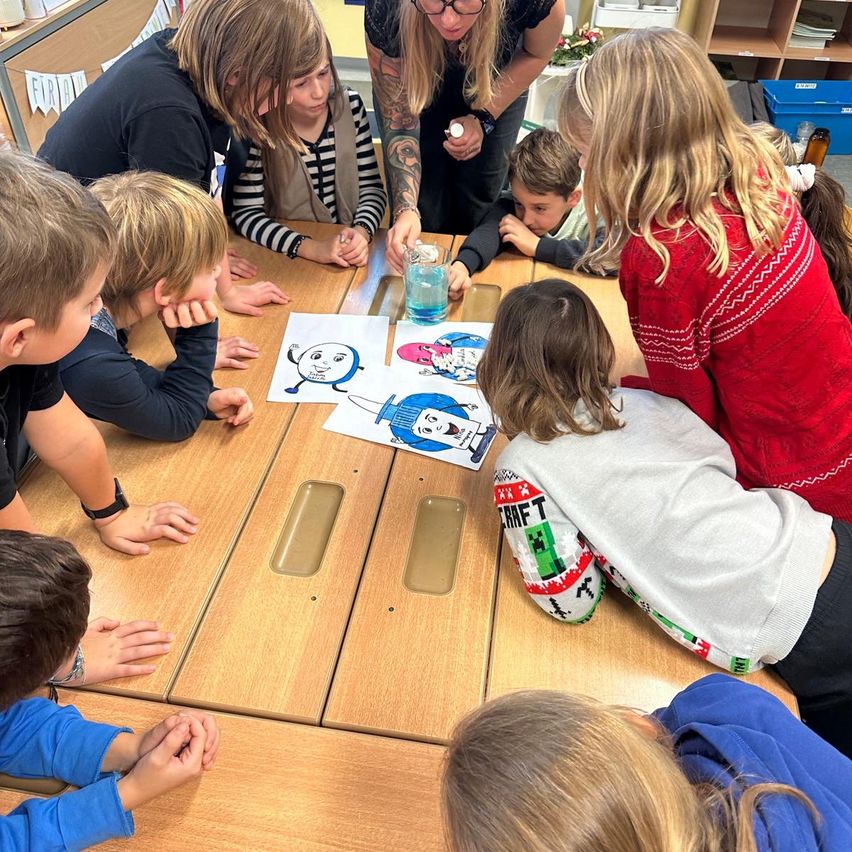 Eine Gruppe von Kindern und zwei Erwachsenen versammelt sich um einen Tisch und betrachtet Malvorlagen. Die Kinder sind engagiert und konzentriert, während die Erwachsenen ihnen helfen.