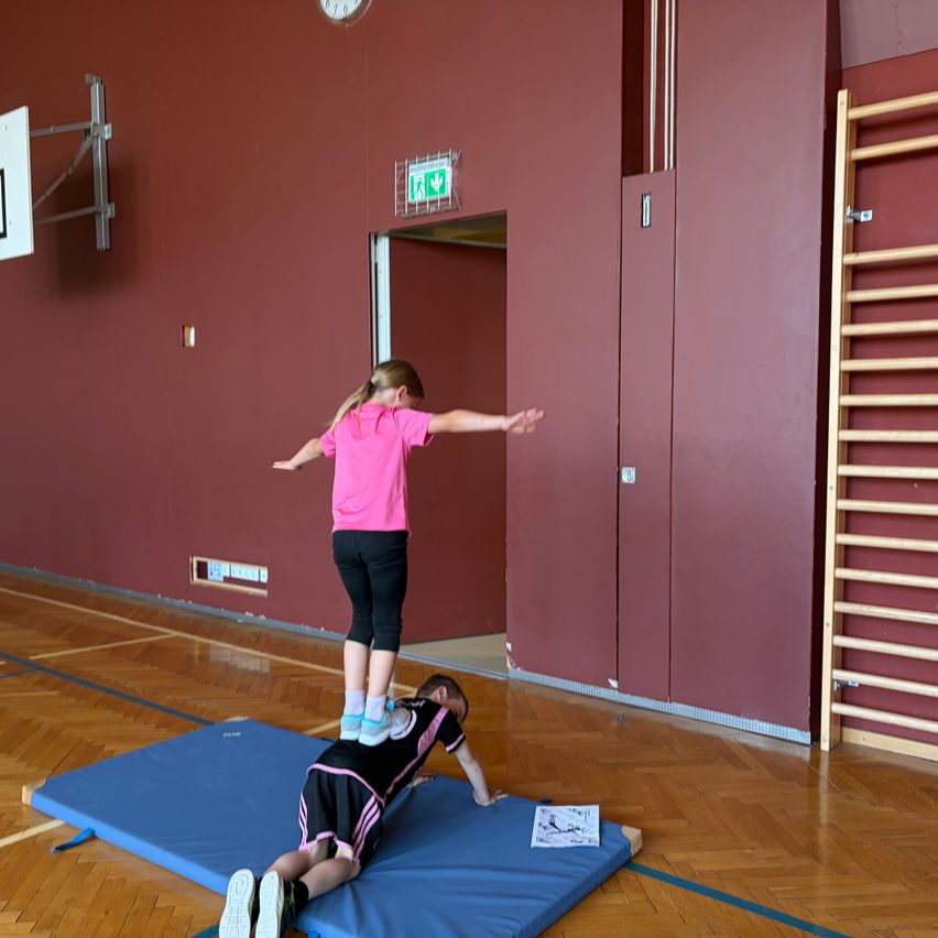 Two children in a gym with one standing on a mat while the other lies down on it, likely practicing gymnastics or martial arts.