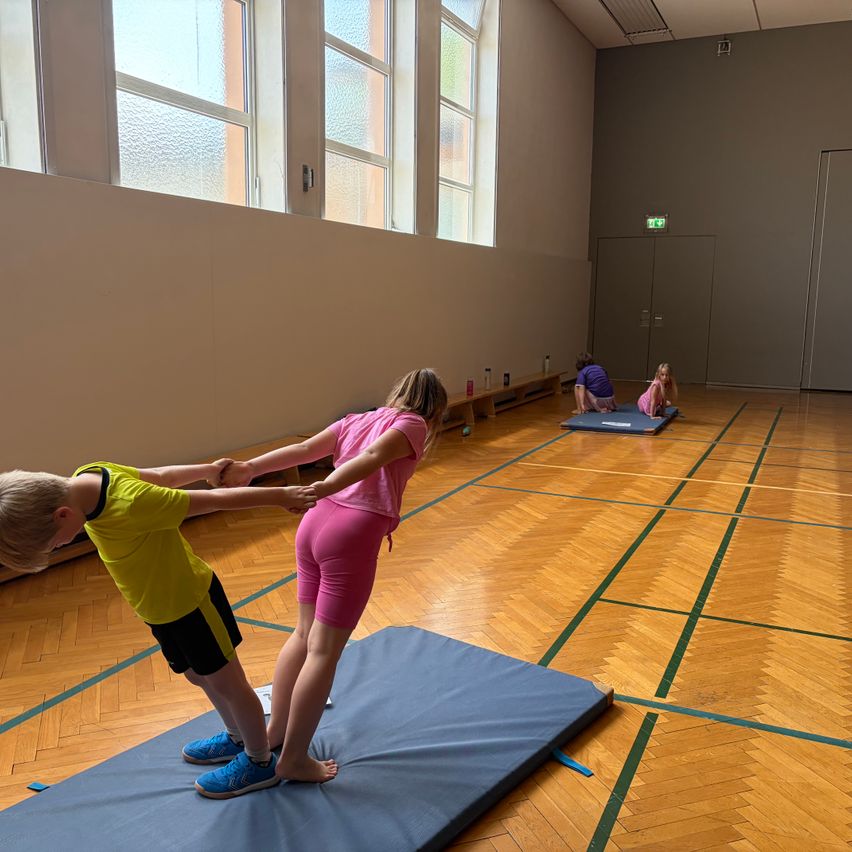 Several children are doing exercises on mats in a gymnasium with large windows and wooden floors. One child is balancing on a mat while holding hands with another.