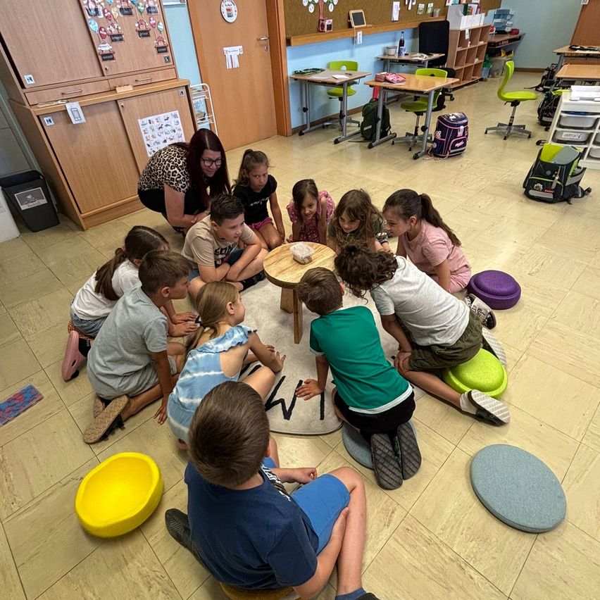 Children sit around a teacher on a floor, listening to a story. The classroom has wooden furniture and colorful bean bags.