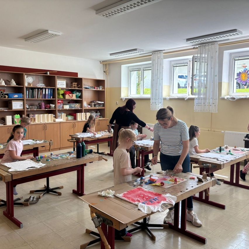 A classroom with wooden desks and chairs. Children work on crafts under the supervision of two women. There is a shelf with various items in the background.