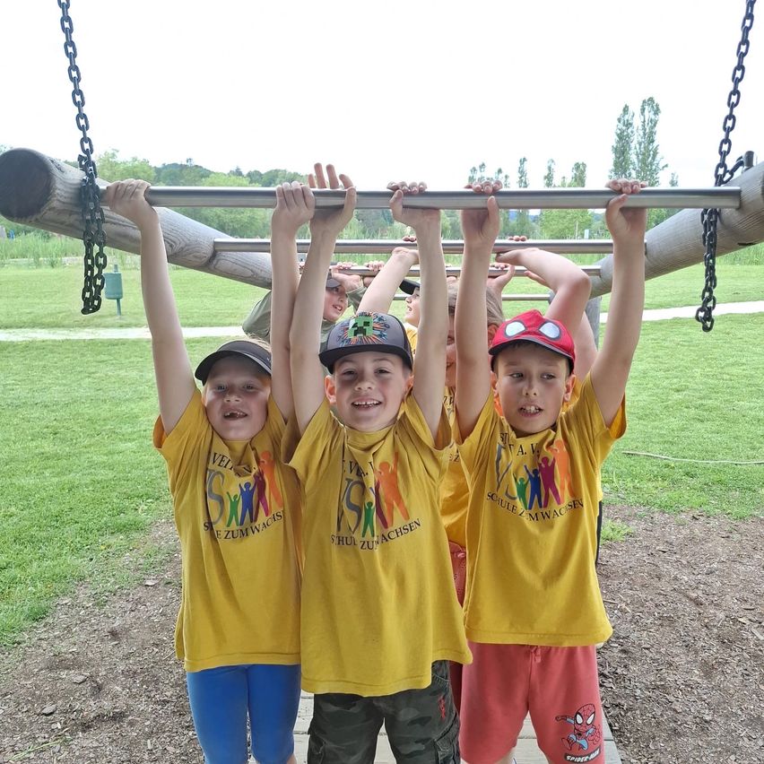 Drei Jungen in gelben T-Shirts mit Designs halten sich an einem horizontalen Balken in einem Park fest. Dahinter sitzt eine Person auf einer Bank. Im Hintergrund sind ein Rasenfeld und Bäume zu sehen.