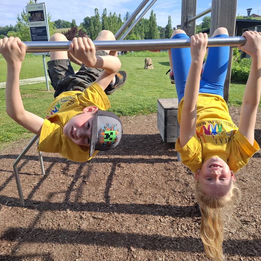Two children in yellow shirts hang upside down on a playground bar, smiling and looking at the camera. A grassy area and a stone structure are visible behind them.