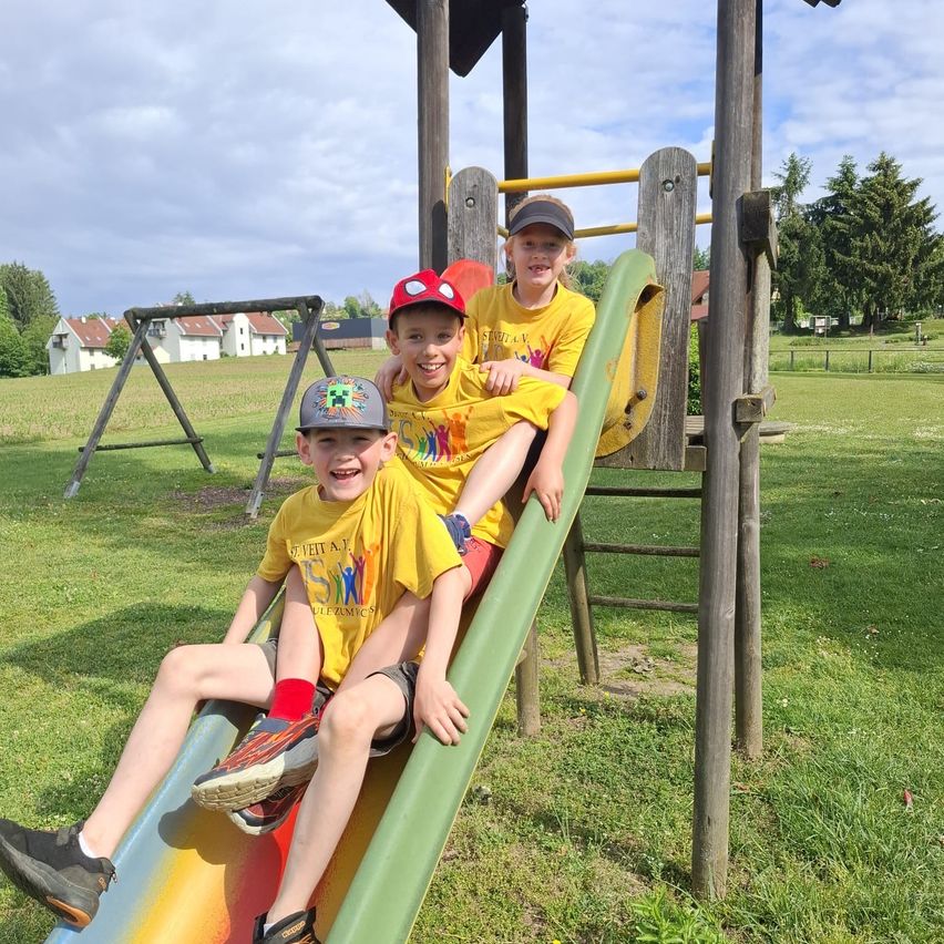Three young boys wearing matching yellow shirts are playing on a colorful slide at a playground, with a swing and houses in the background.