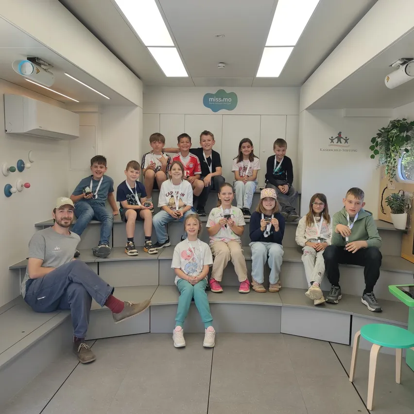 Eine Gruppe von Kindern und ein erwachsener Mann sitzen auf einer Bank in einem Raum mit weißen Wänden und Deckenbeleuchtung.