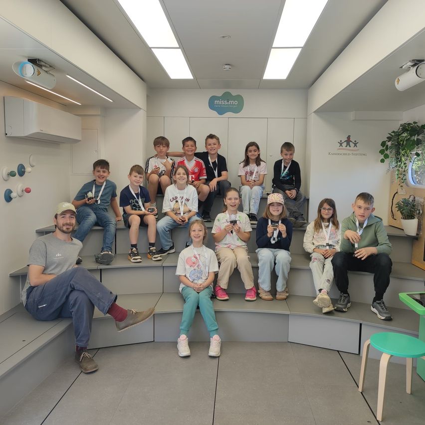Eine Gruppe von Kindern und ein erwachsener Mann sitzen auf einer Bank in einem Raum mit weißen Wänden und Deckenbeleuchtung.