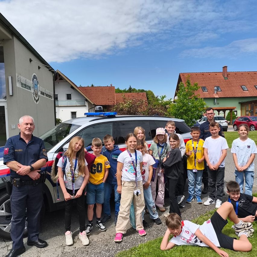 A group of children and two police officers pose in front of a police car. They stand on the grass with houses and trees in the background.